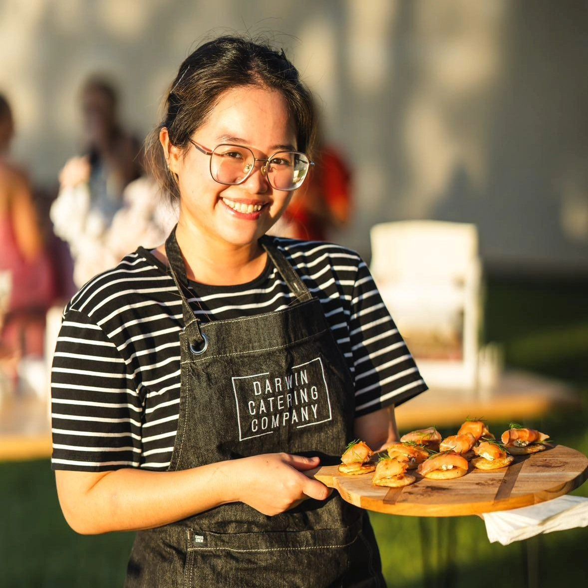 A Woman Wearing an Apron That Says Darryn Catering Company is Holding a Tray of Food — Darwin Catering Company in Brinkin, NT