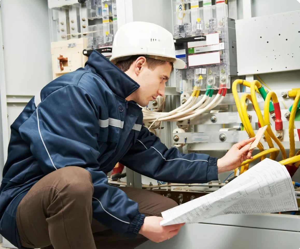 Electrician in a blue jacket examines wiring diagram at electrical panel.