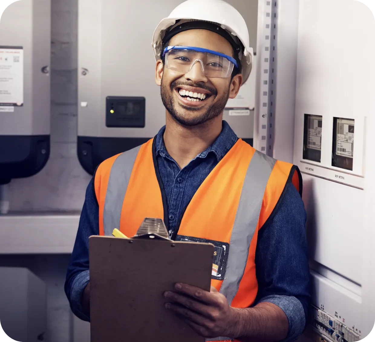 Smiling electrician in safety gear holding a clipboard in an electrical room.