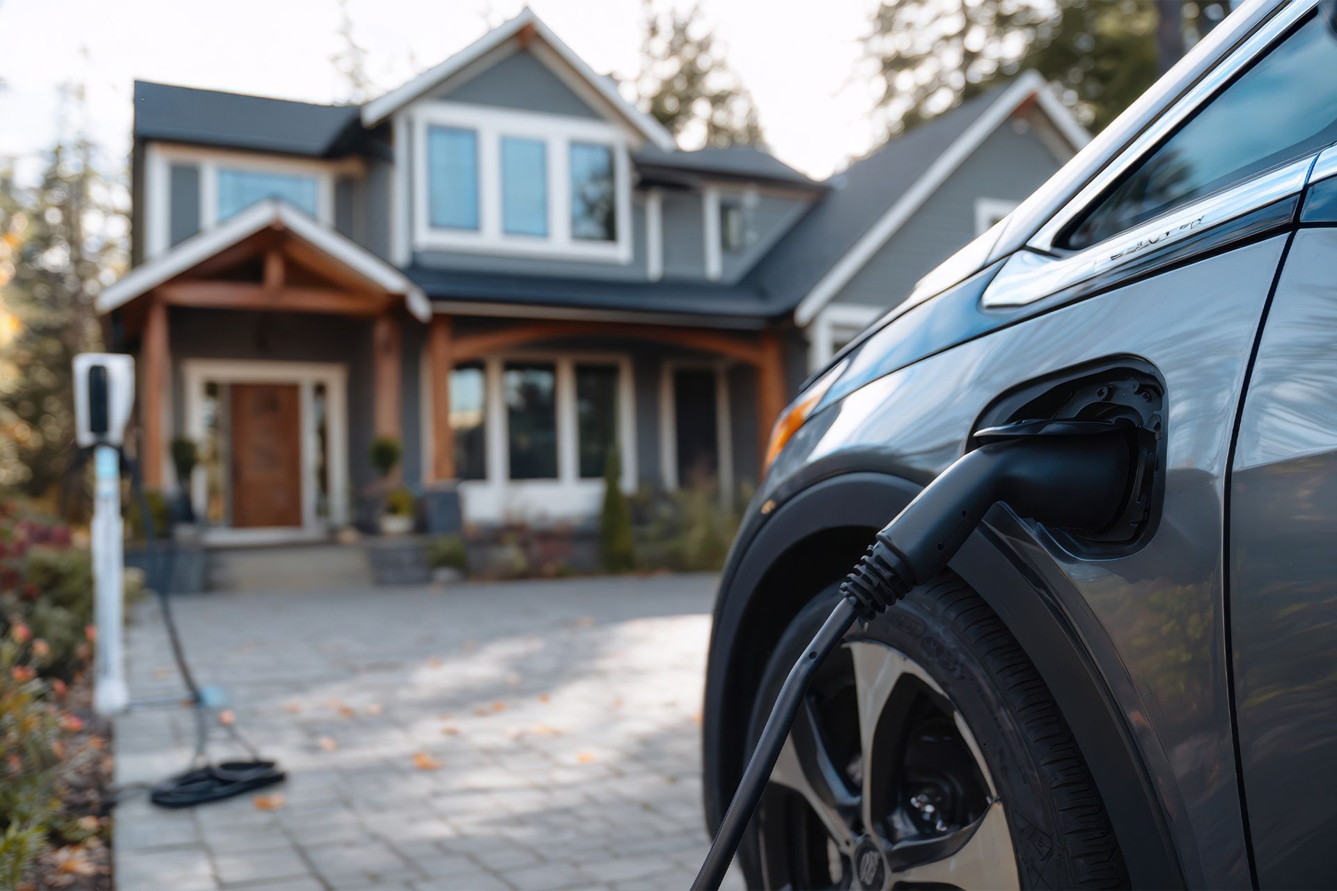 Electric car charging at a residential home. Grey car, black charging cable. House with gray siding.
