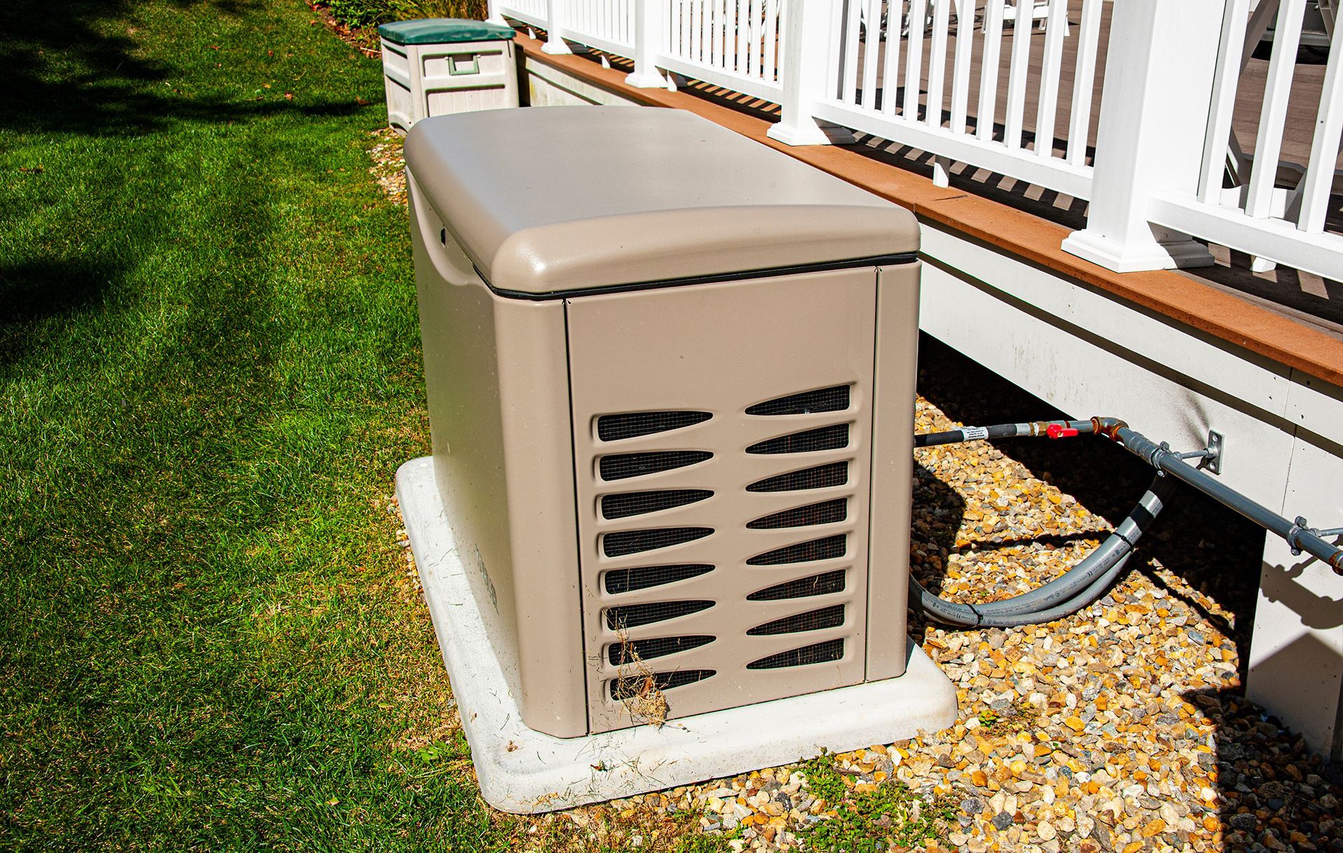 Tan outdoor generator next to a house with a white railing. The generator sits on a white pad on gravel.
