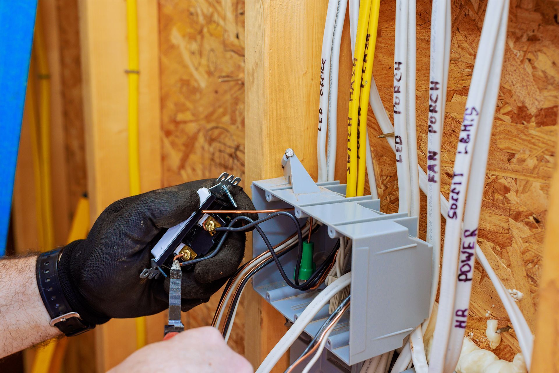 Electrician wiring cables inside a wall box; black gloves, wire cutter, wires labeled