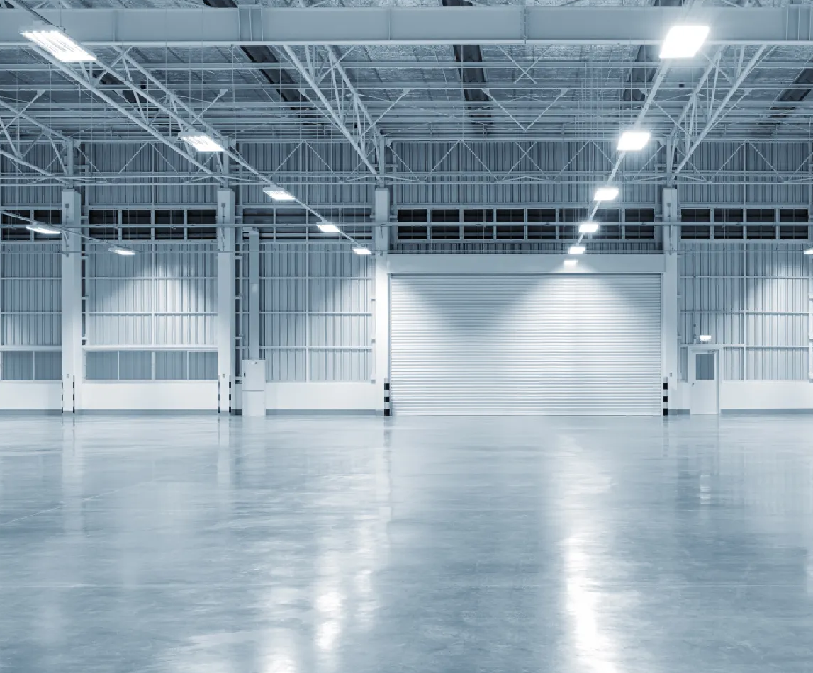 Empty, industrial warehouse interior; concrete floor reflects overhead lights, open bay door.