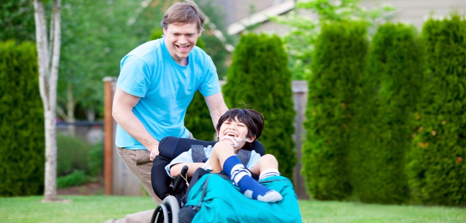 Uomo che spinge un bambino sorridente su una sedia a rotelle sull'erba; sono all'aperto vicino a cespugli verdi.