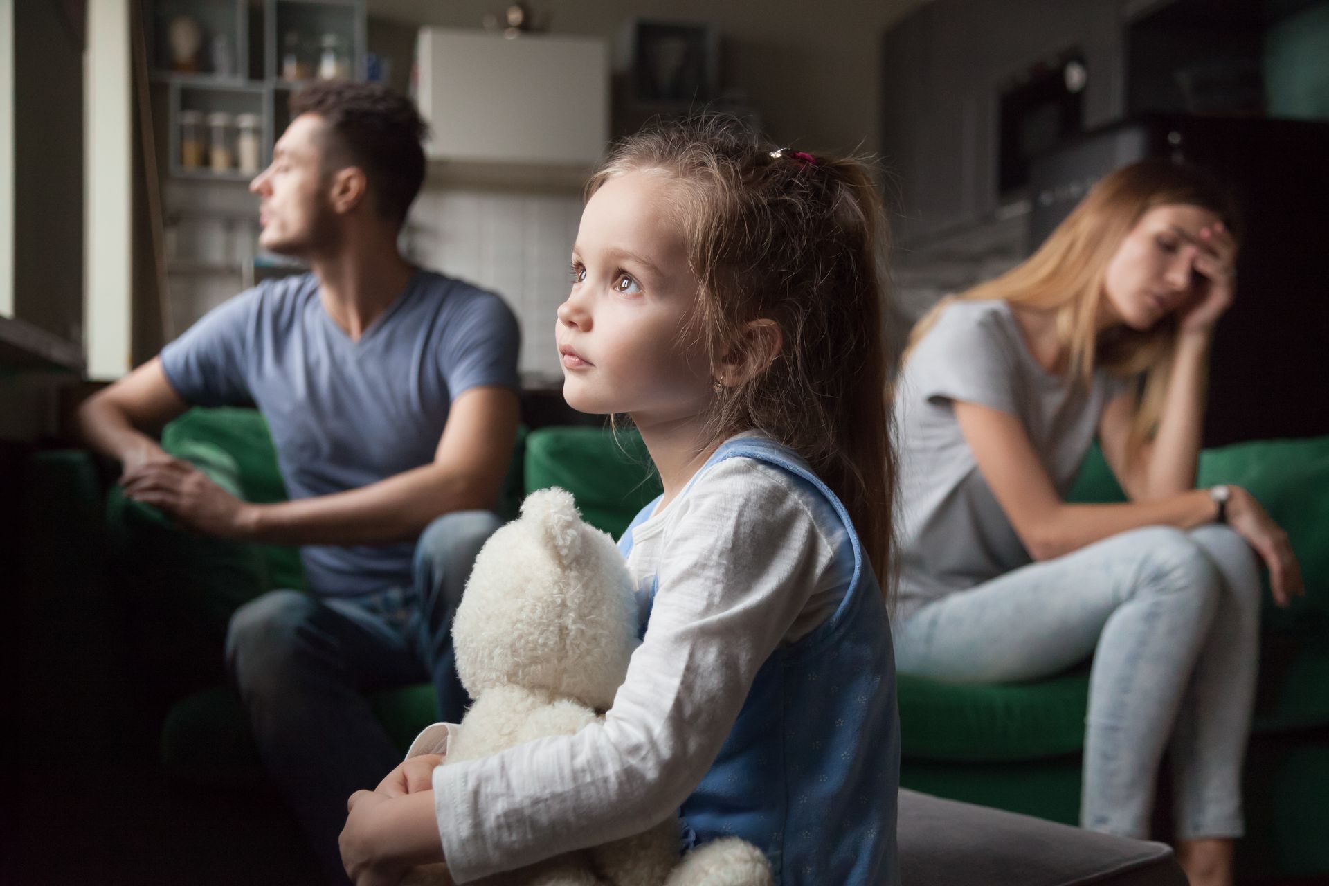 Bambina che tiene in mano un orsacchiotto, guardando in alto. Genitori sullo sfondo, separati