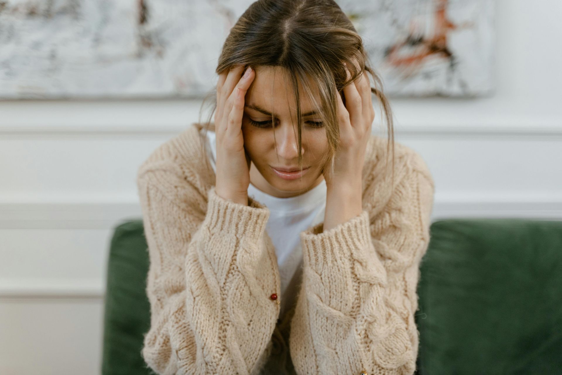 Donna con le mani sulla testa, che guarda in basso, con un maglione in tonalità neutre seduta su un divano verde.