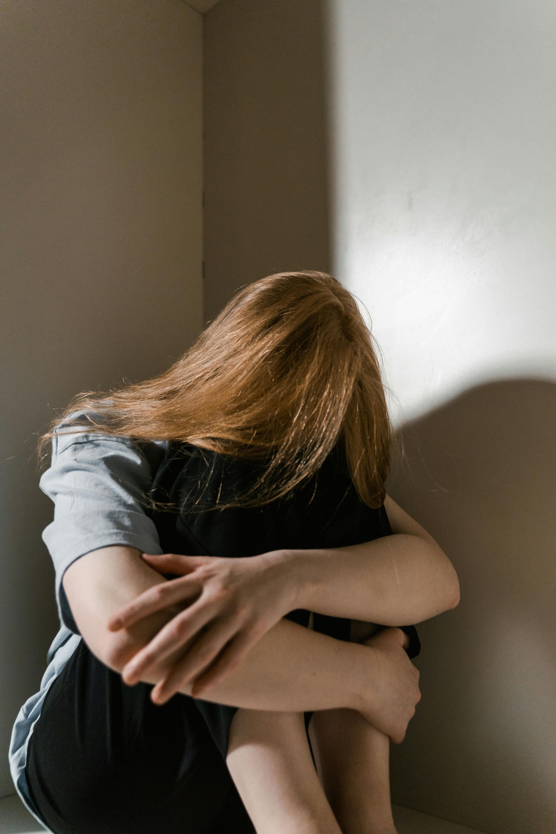 Donna con i capelli rossi, seduta in un angolo, con la testa bassa e le braccia attorno alle gambe.