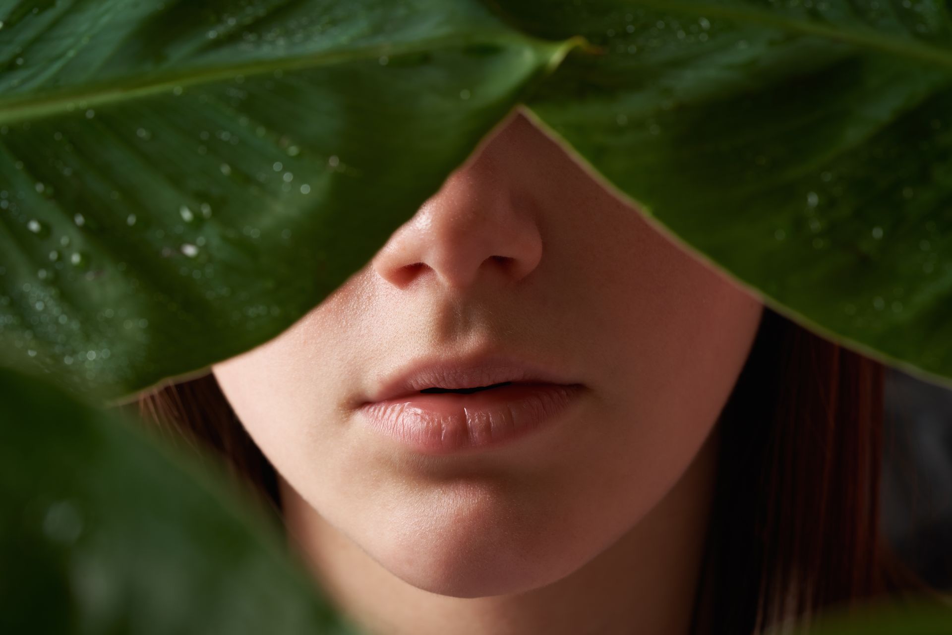 Primo piano del viso di una persona, con naso e bocca incorniciati da grandi foglie verdi.