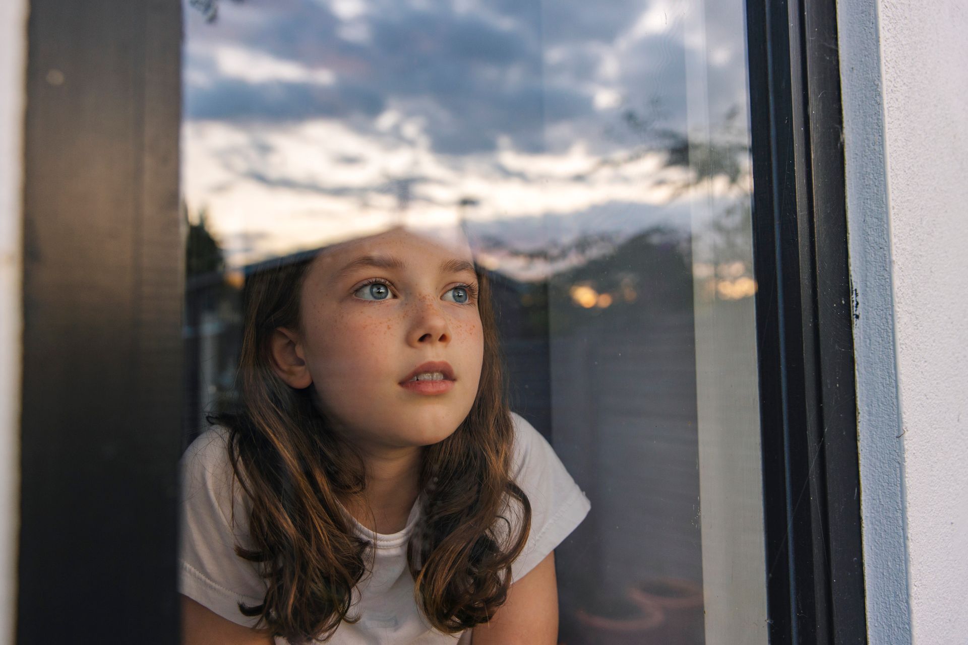 Ragazza che guarda fuori da una finestra al tramonto, il cielo riflesso nel vetro.