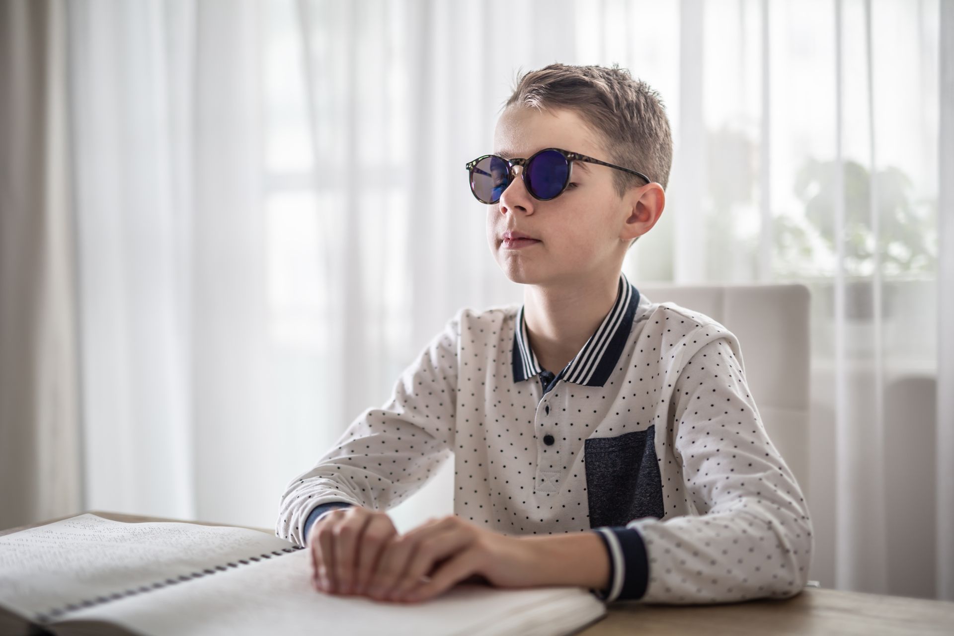 Ragazzo con occhiali da sole che legge il braille, seduto a un tavolo davanti a una finestra.