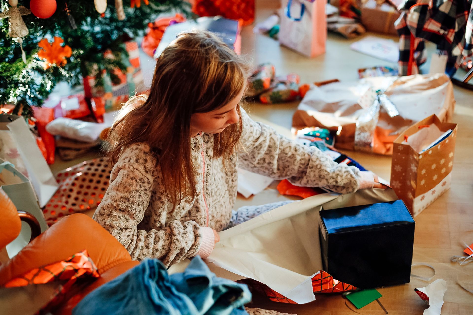 Ragazza che scarta regali sul pavimento vicino a un albero di Natale; circondata da regali e carta da regalo.