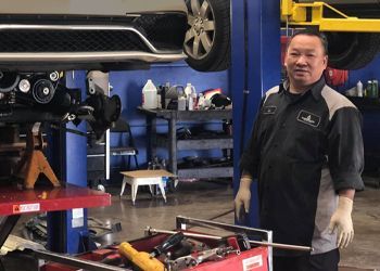 A man is standing in front of a car on a lift in a garage.