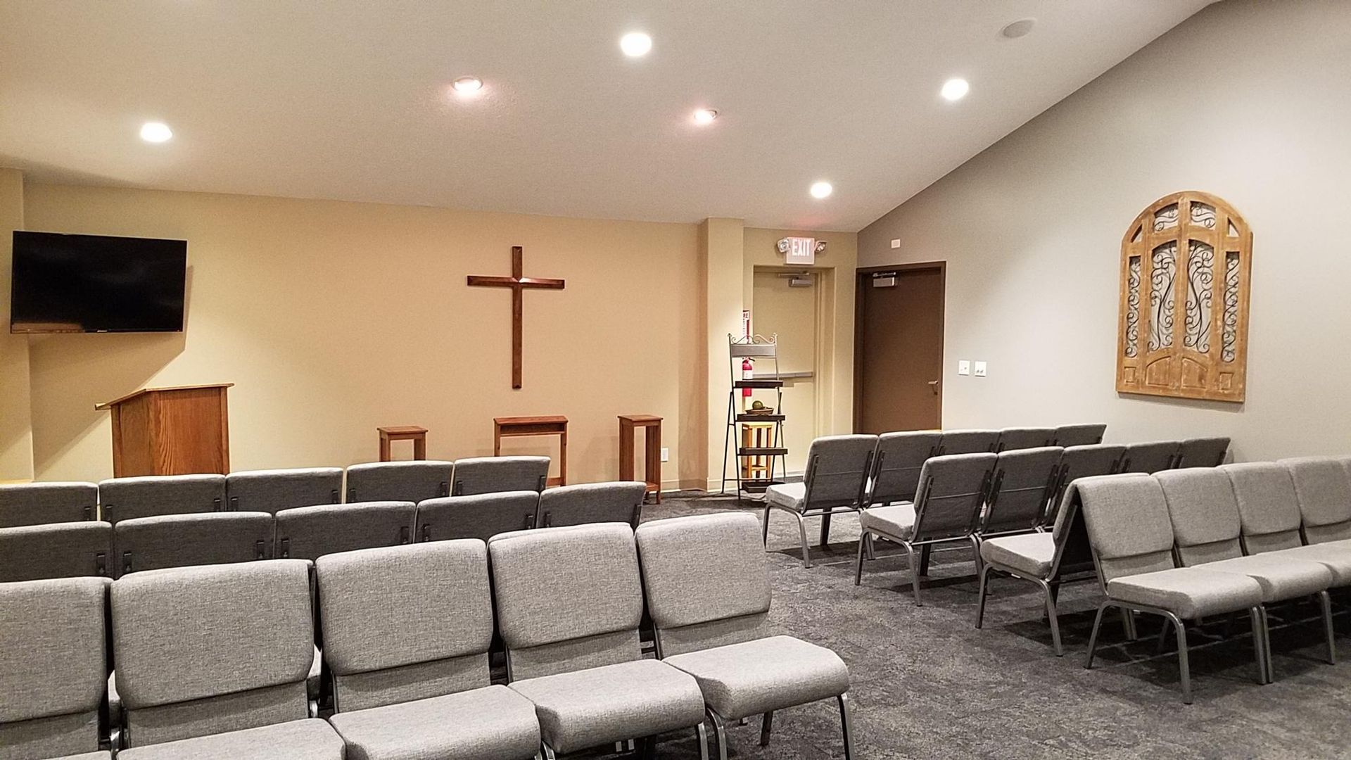 Interior of a chapel with rows of gray seating, a wooden cross, and an ornate wall carving.