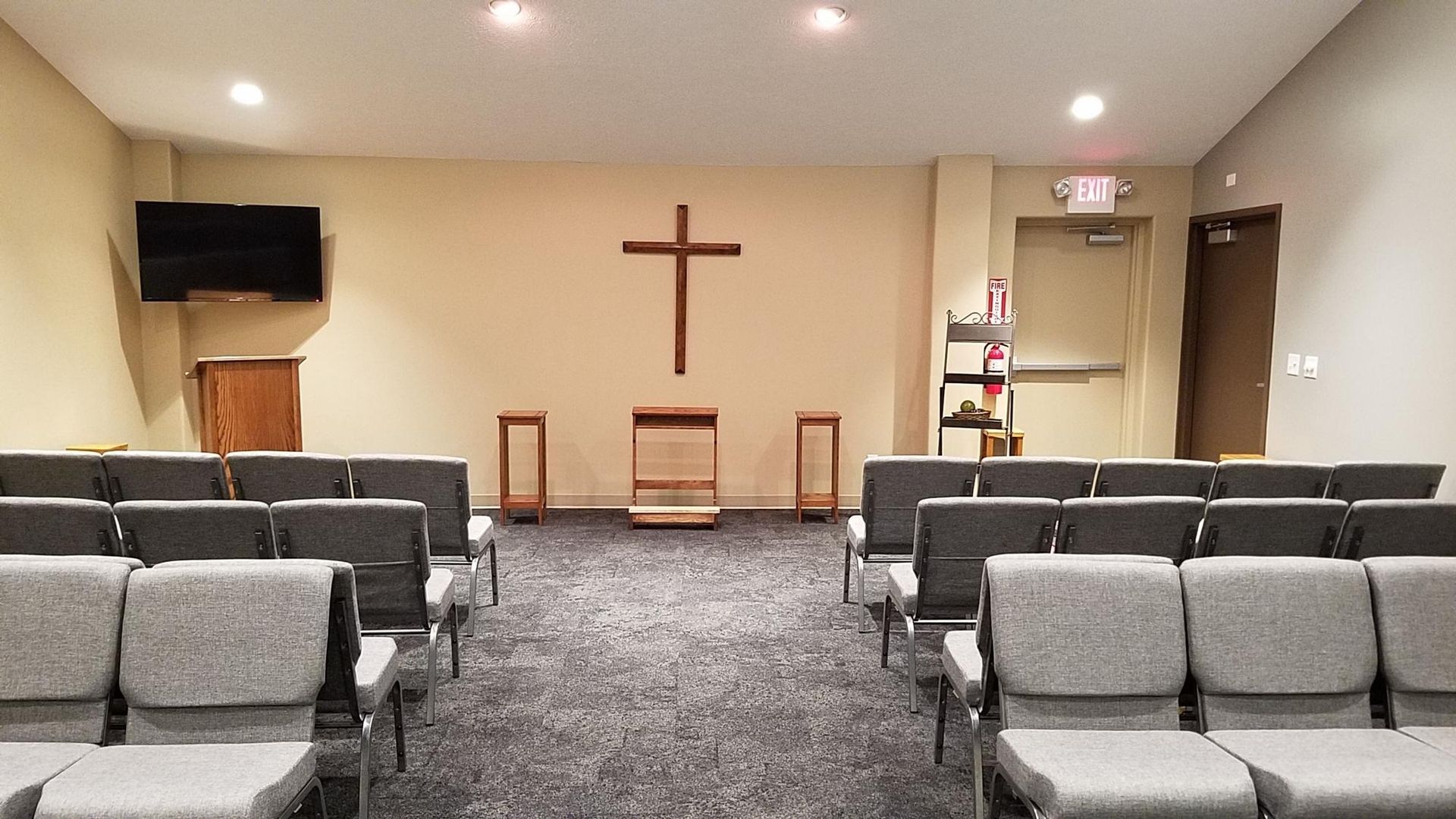 Rows of gray chairs face a wooden cross on the wall of a chapel; exit door visible.