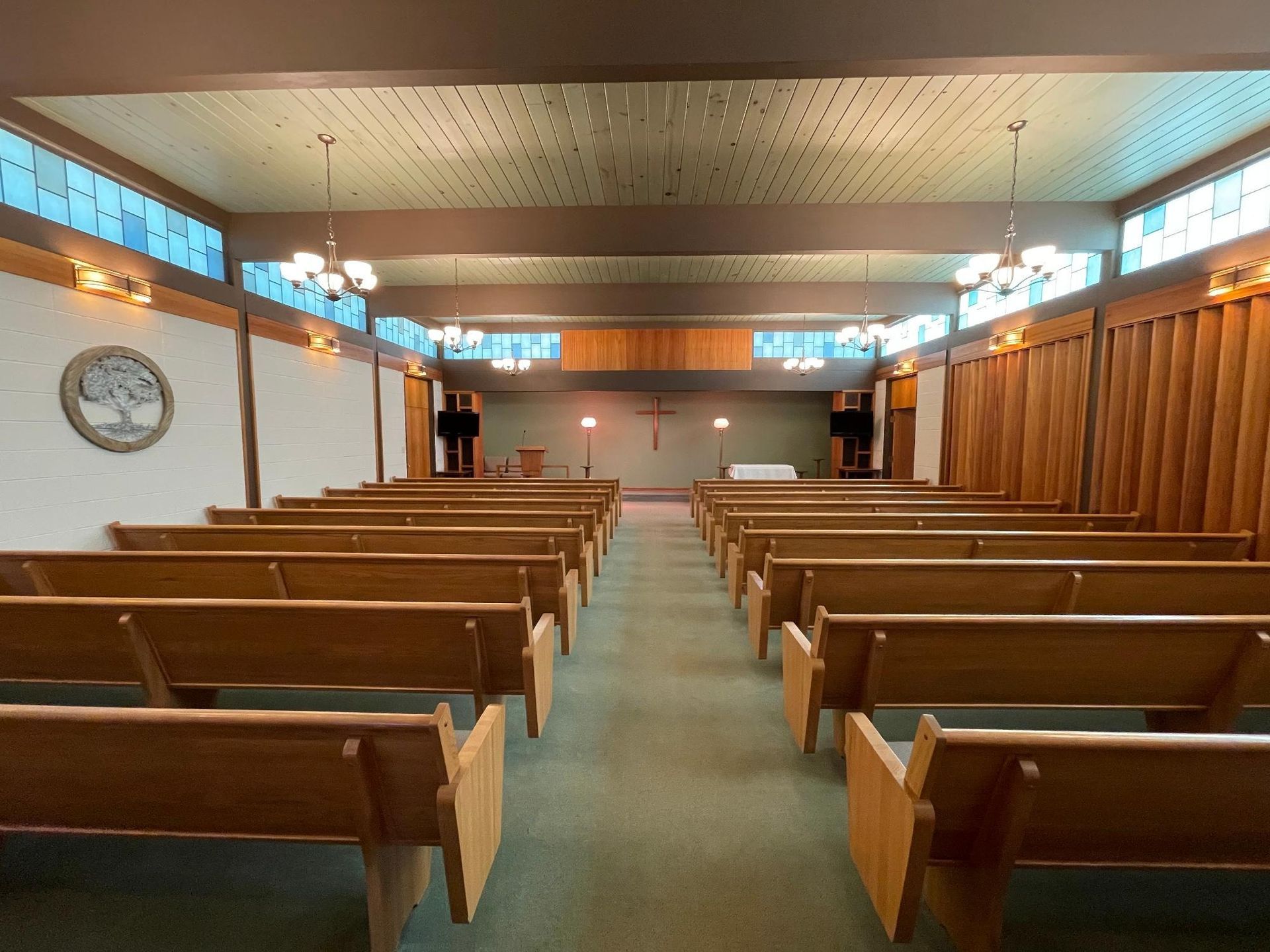 Interior view of a chapel with wooden pews, a green carpet, and a cross at the altar.