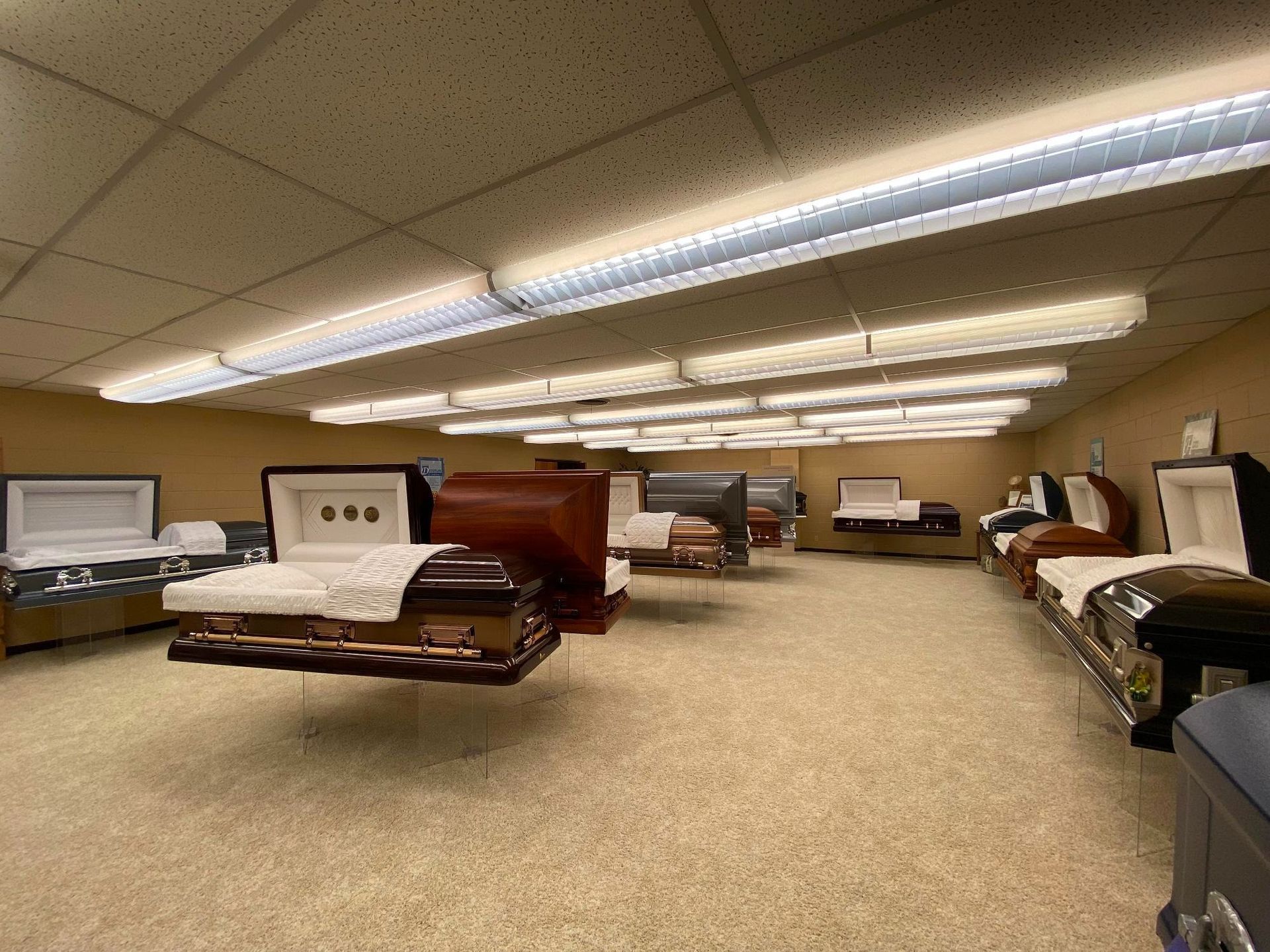 Interior of a funeral home displaying several open coffins on display. Room is beige, lit by fluorescent lights.
