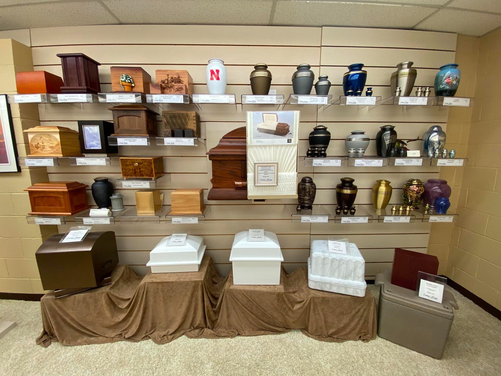 Display of urns and caskets on shelves in a funeral home. Various sizes, colors, and designs are shown.