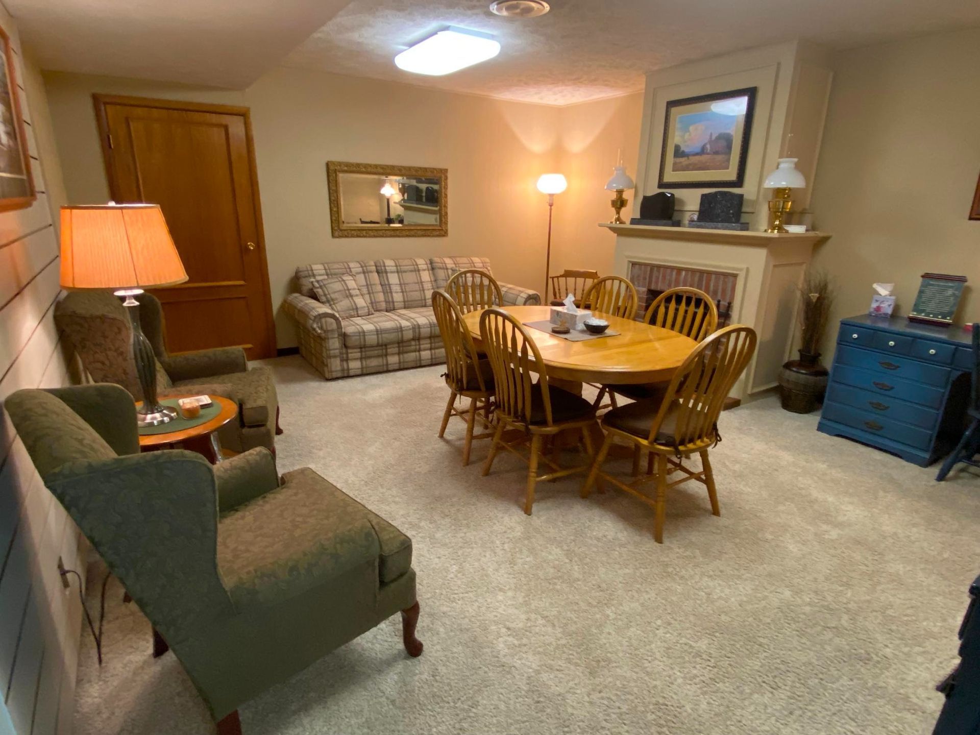 Cozy living room with seating, a dining table, and a fireplace. Beige carpet, soft lighting, and neutral-colored walls.