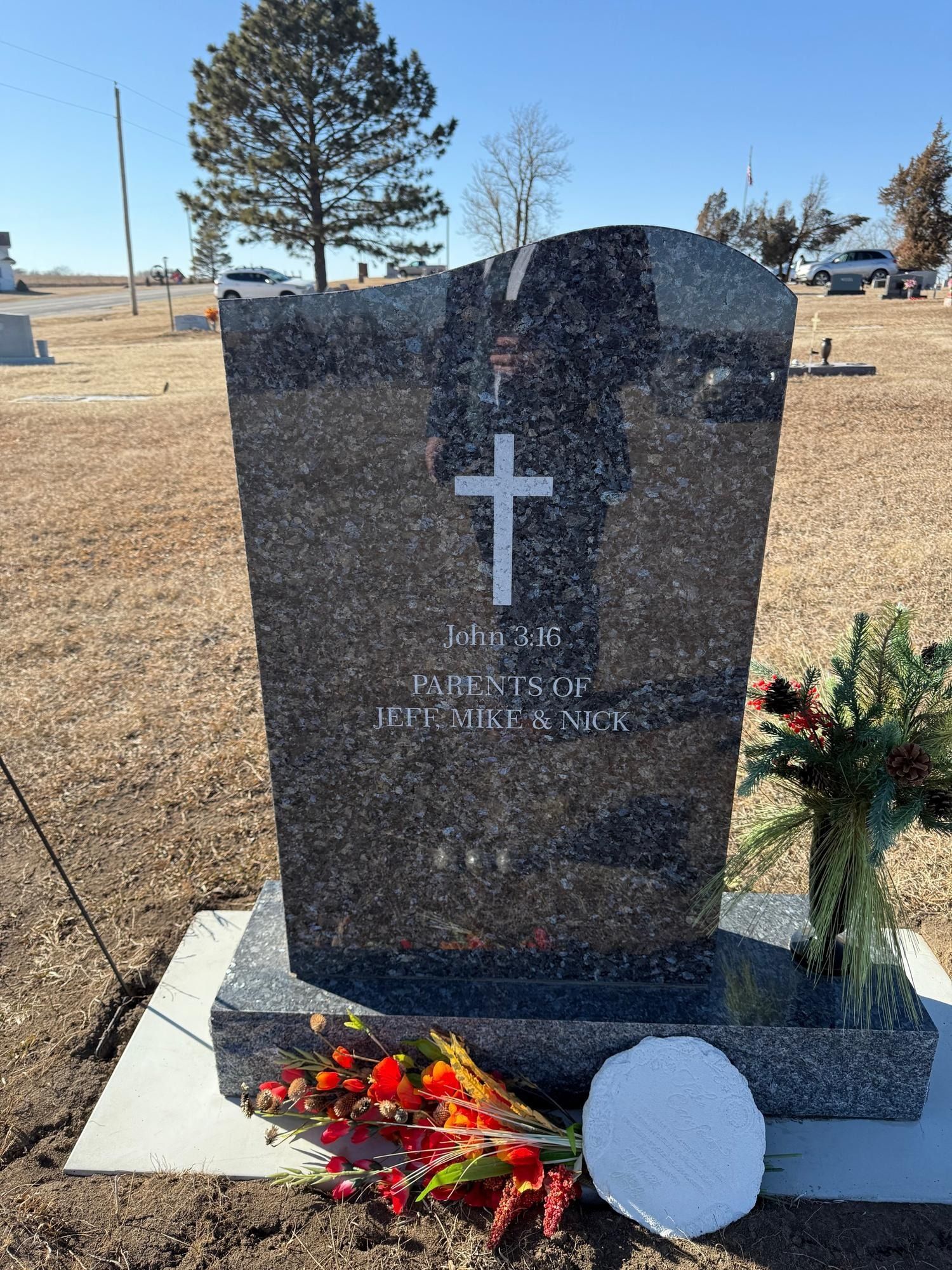 Grave marker with a cross and inscription, with flowers at its base, set in a cemetery.