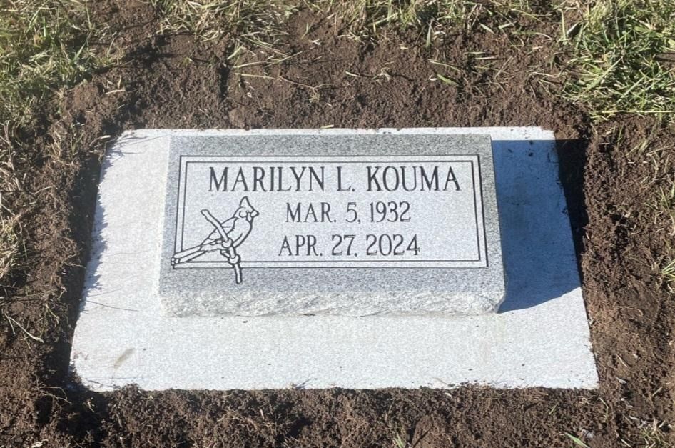 Gray granite headstone with inscription for Marilyn L. Kouma, born 1932, died 2024, featuring a bird and a floral motif.