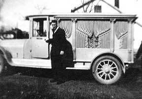Man standing by an old hearse. Black suit. Outdoor setting with house visible.