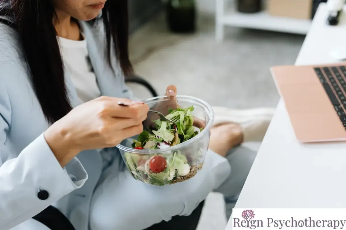 A person wearing a light blue blazer eats a green salad from a glass bowl while working at a desk with a laptop.