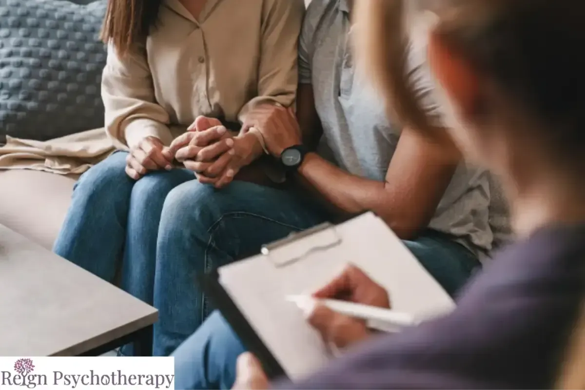 A couple sitting on a couch and holding hands while a therapist taking notes.