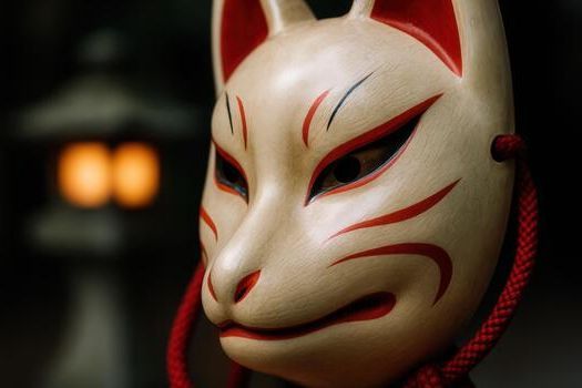 Fox mask, off-white with red markings, red cord, in front of a blurred lantern.