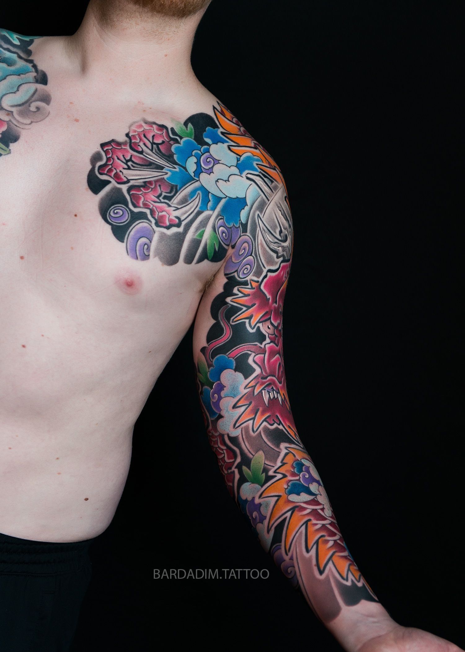 Man's torso and arm with a colorful, detailed Japanese-style floral tattoo sleeve against a black backdrop.
