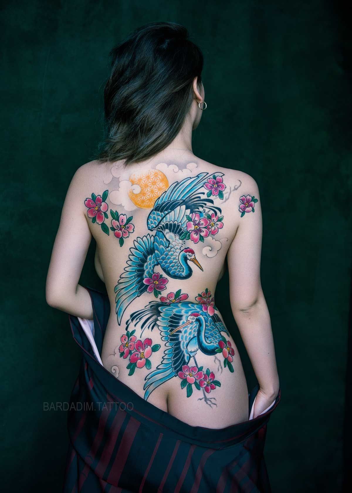 Woman with back tattoo of two blue cranes, pink flowers, and a yellow sun; standing against a green backdrop.