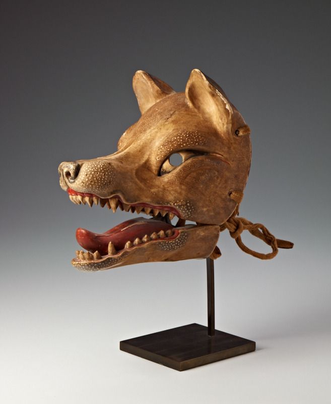 A carved wooden Kitsune fox mask with an open jaw, red tongue, and tan fur. Mounted on a black stand.