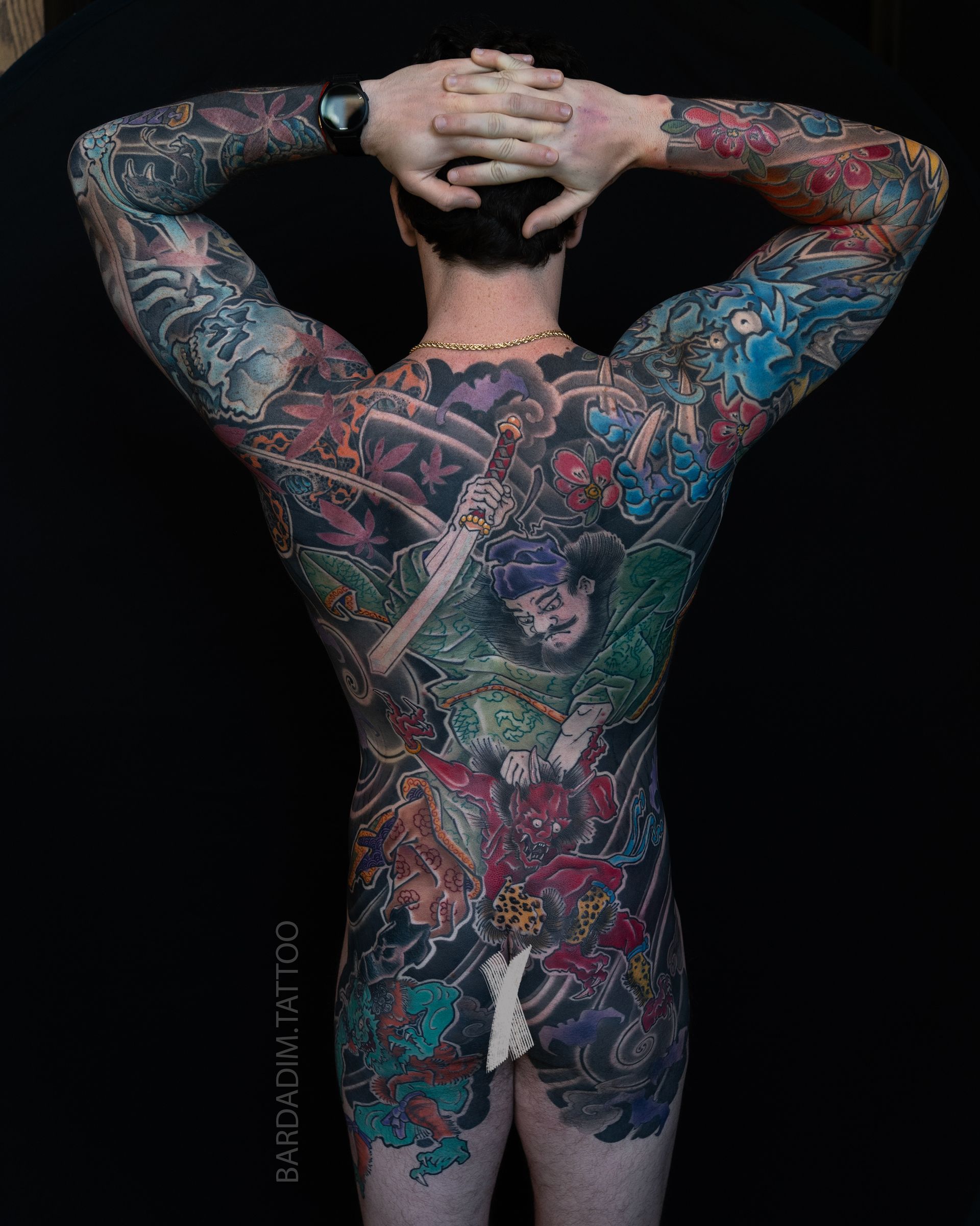 Person with full-body colorful tattoos, back to camera, hands behind head. Dark background at Bardadim Tattoo.