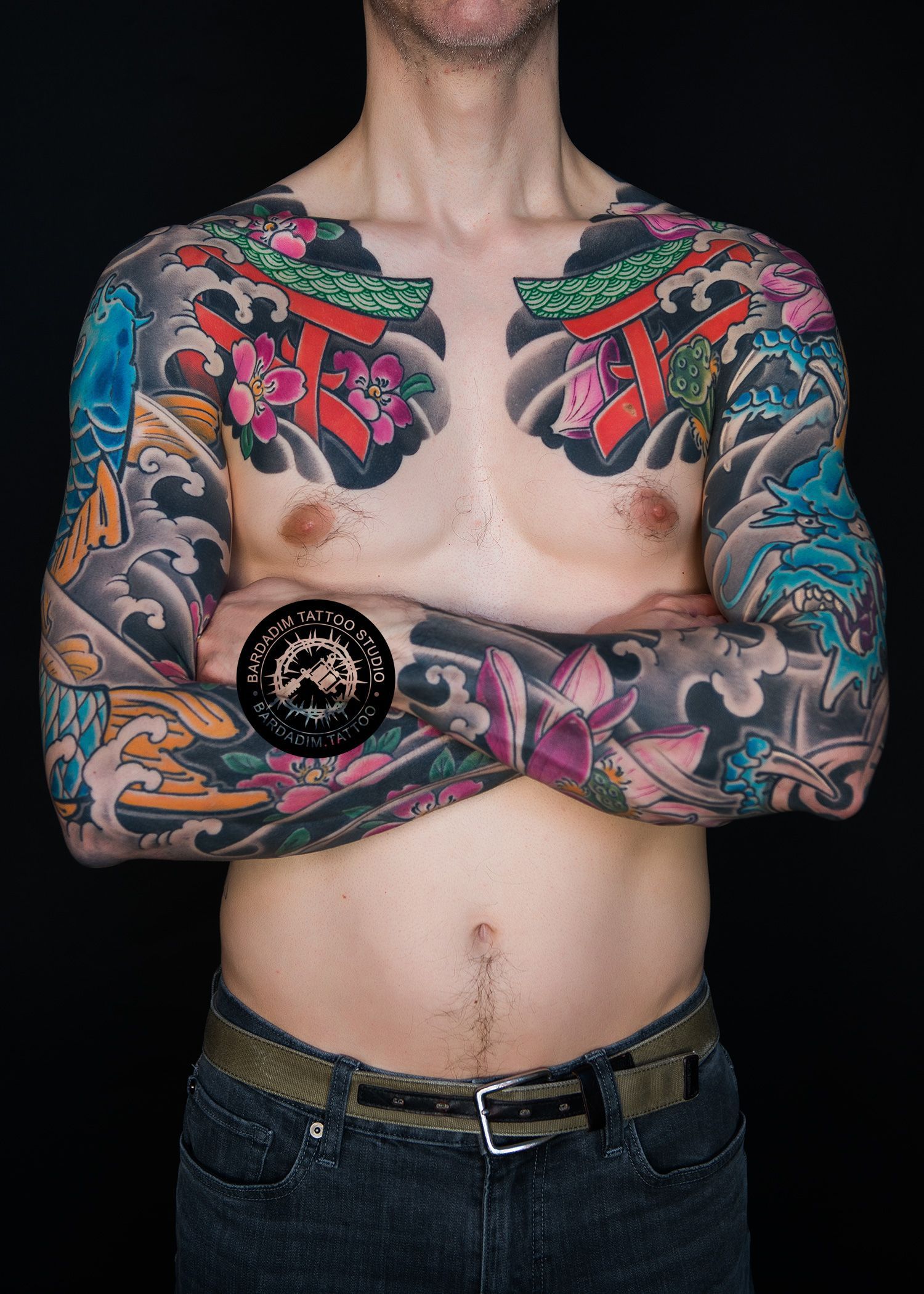 Man with full sleeve tattoos, arms crossed, torso partially visible against black background.