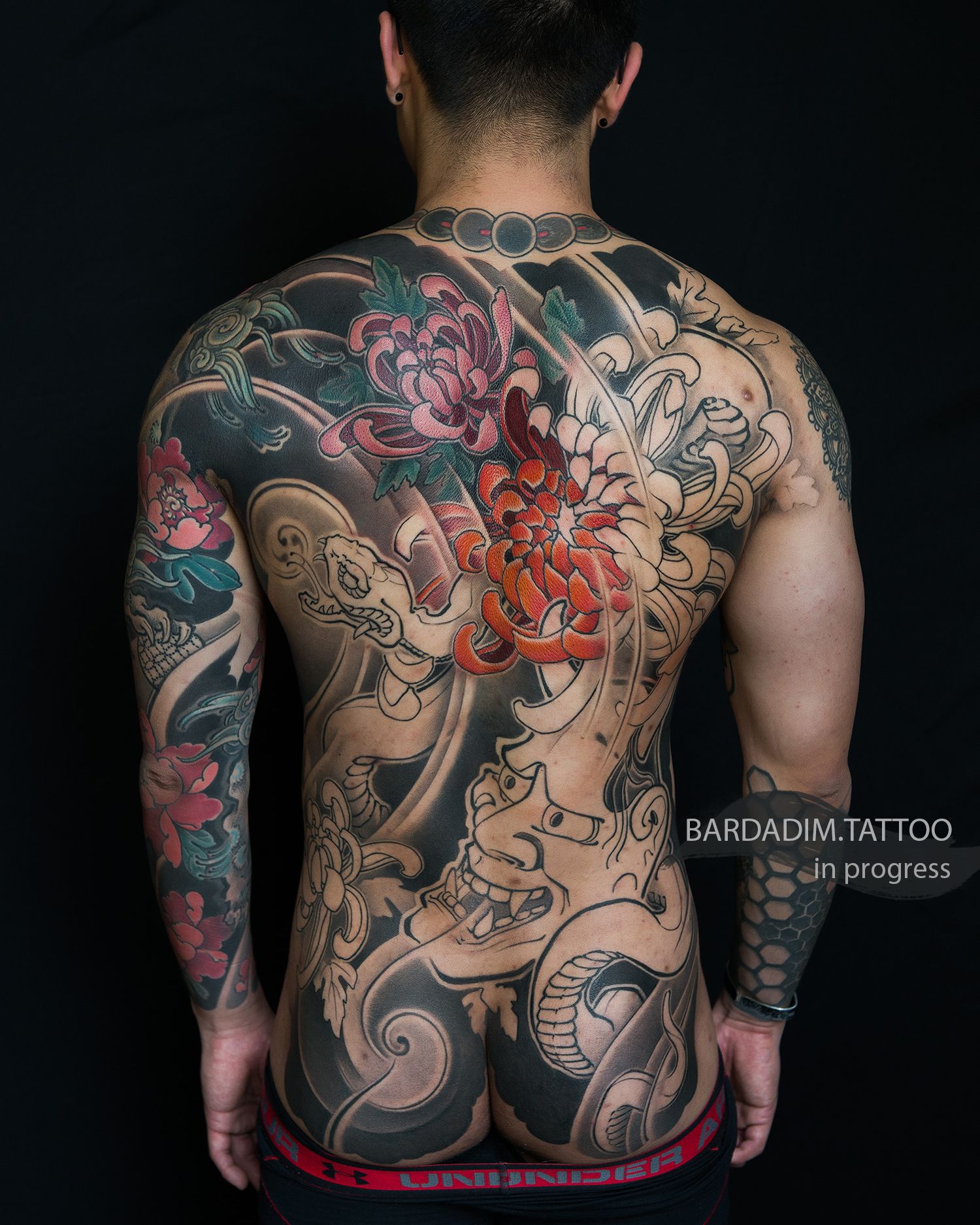 Full back tattoo in progress, featuring Japanese floral and wave motifs in black, red, and teal against dark skin.