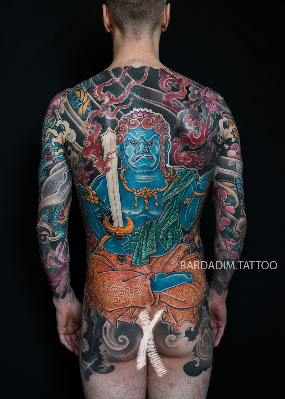 Man's back covered in a large, colorful Japanese-style tattoo featuring a blue deity Fudo-Myoo holding a sword.