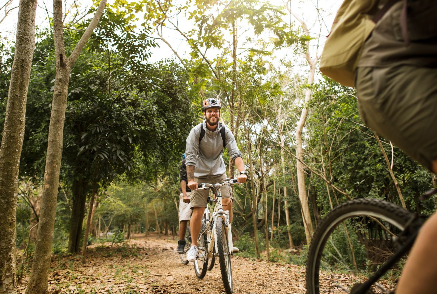 Three people riding bikes on a dirt path in a sunlit forest.