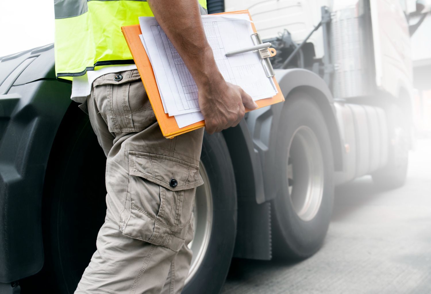 Person in reflective vest holding clipboard, inspecting a semi-truck's tire in a bright, outdoor setting.