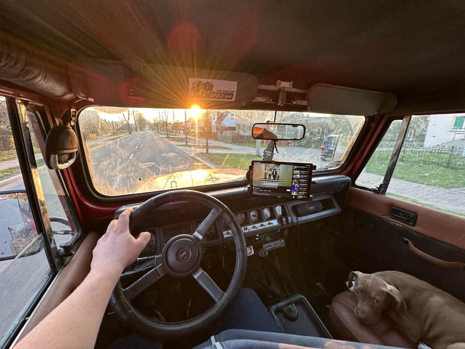 Inside a vintage red Jeep Wrangler, a driver holds the steering wheel while a dog sits in the passenger seat; sunset outside.