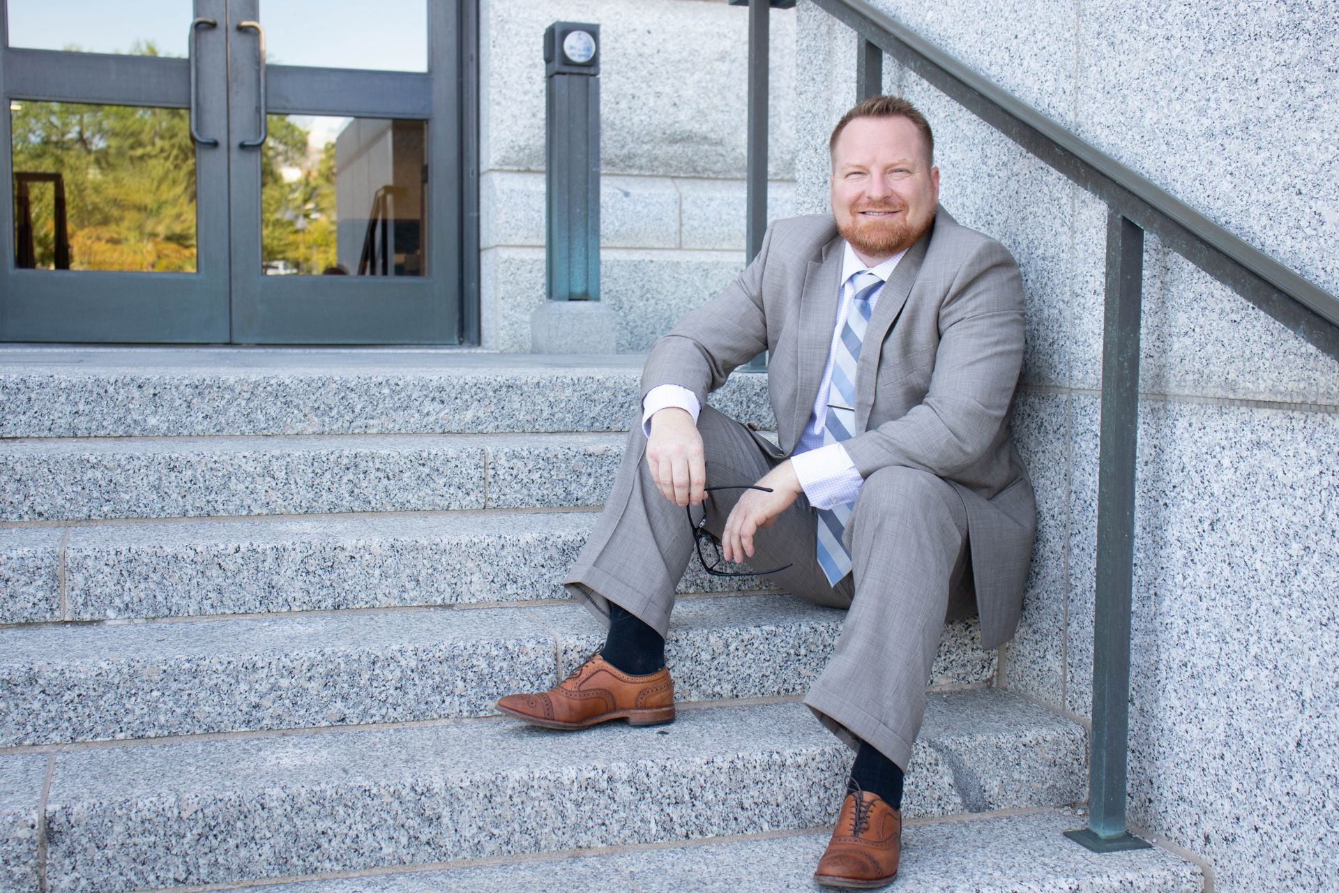 Man in suit sits on stone steps, smiling. Door behind him, outdoors.