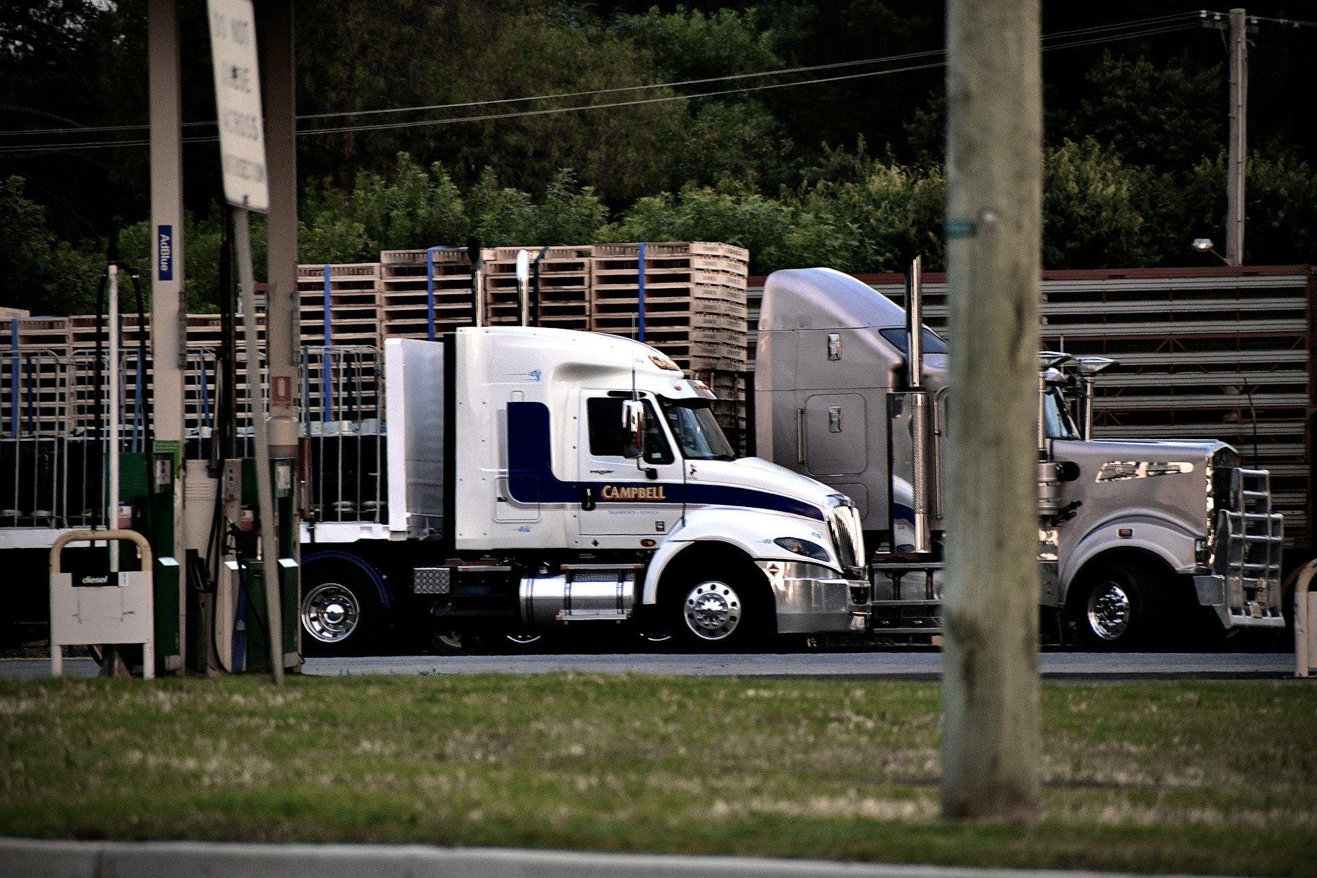 Two semi-trucks parked at a loading dock. One is white with blue stripes, the other tan.