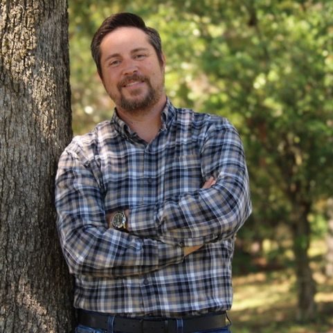 Man with crossed arms leaning against a tree, smiling. Wearing plaid shirt, watch, and jeans. Outdoors.