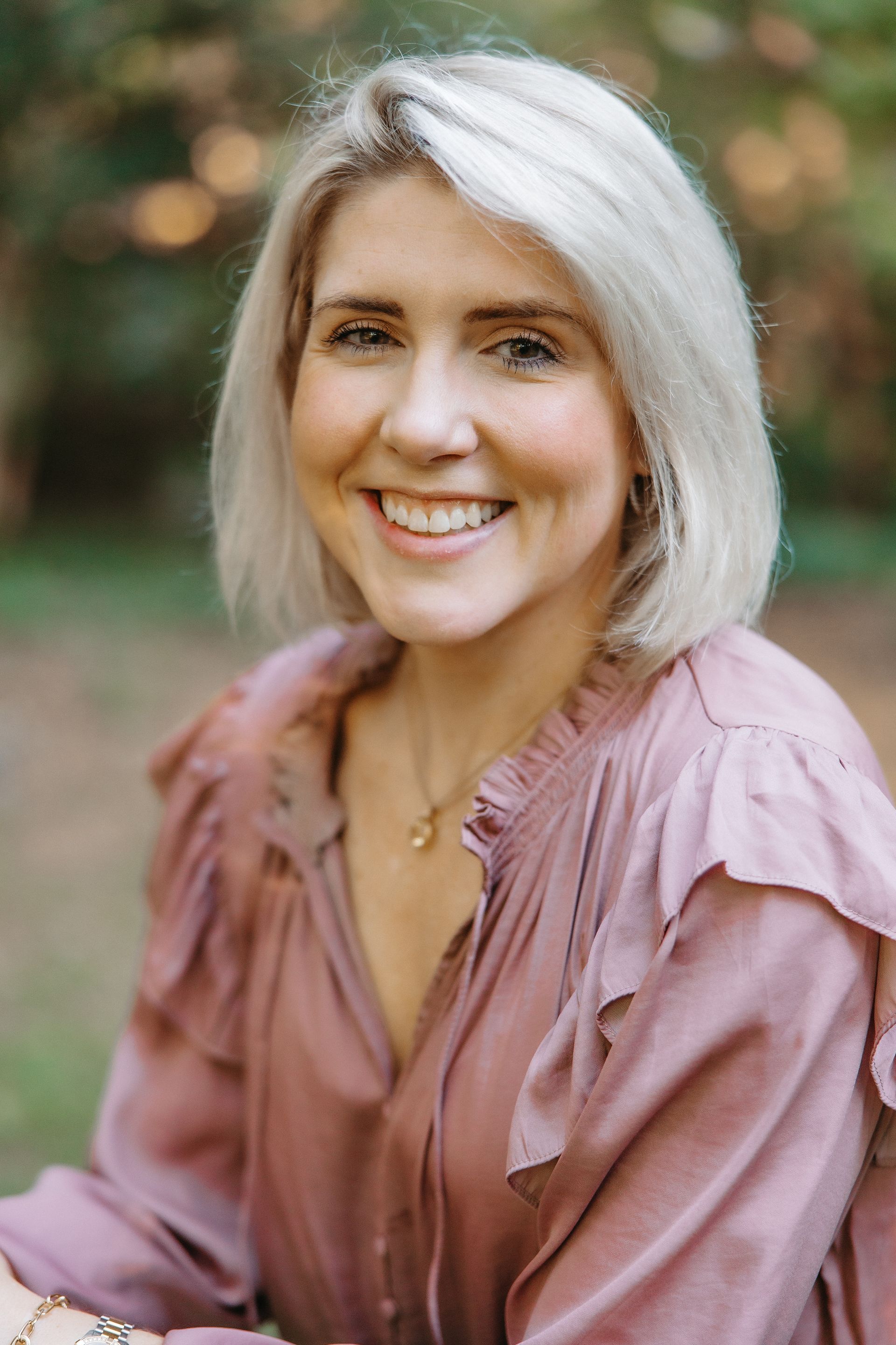 Blonde woman smiles, wearing a pink ruffled shirt, outdoors.