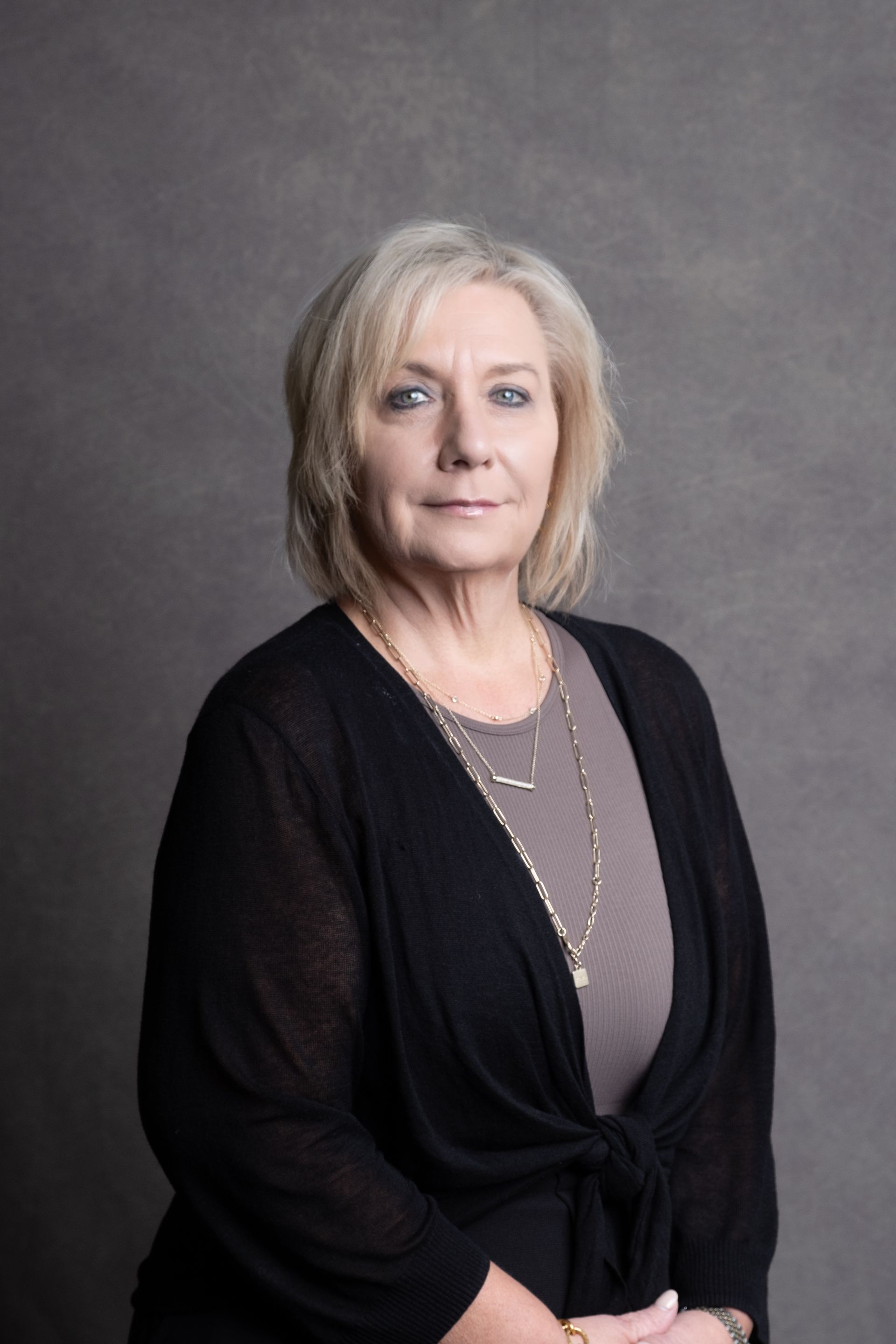 Woman with blonde hair, wearing black cardigan, standing against gray backdrop.