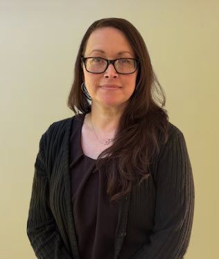 Woman with glasses wearing a black cardigan, smiling, facing the camera against a light yellow background.
