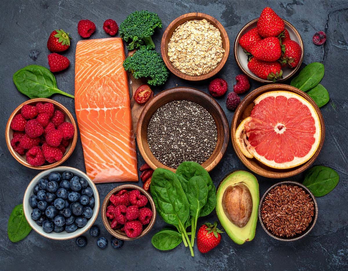 A variety of healthy whole foods, including salmon, berries, broccoli, avocado, grains, and chia seeds on a dark surface.