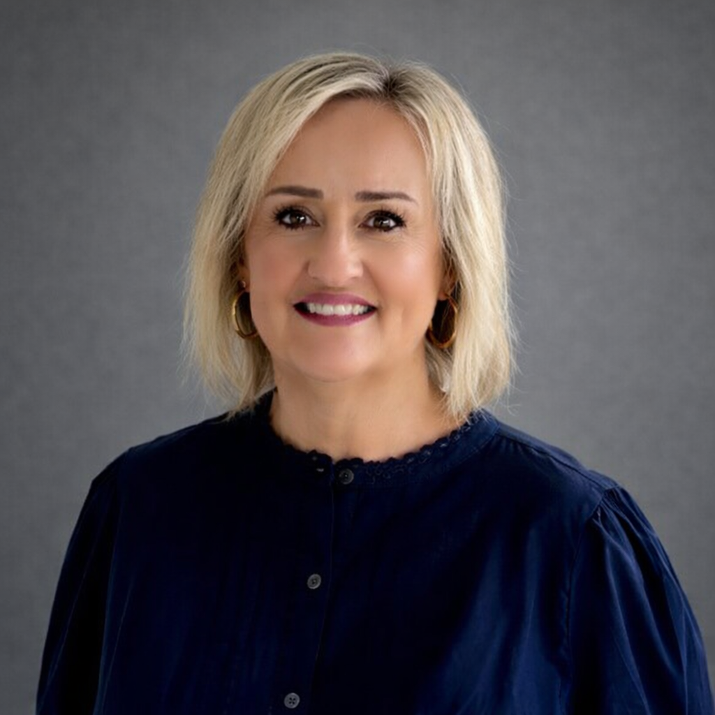 A smiling woman with blonde hair, wearing a dark blue button-down shirt, against a plain gray studio background.