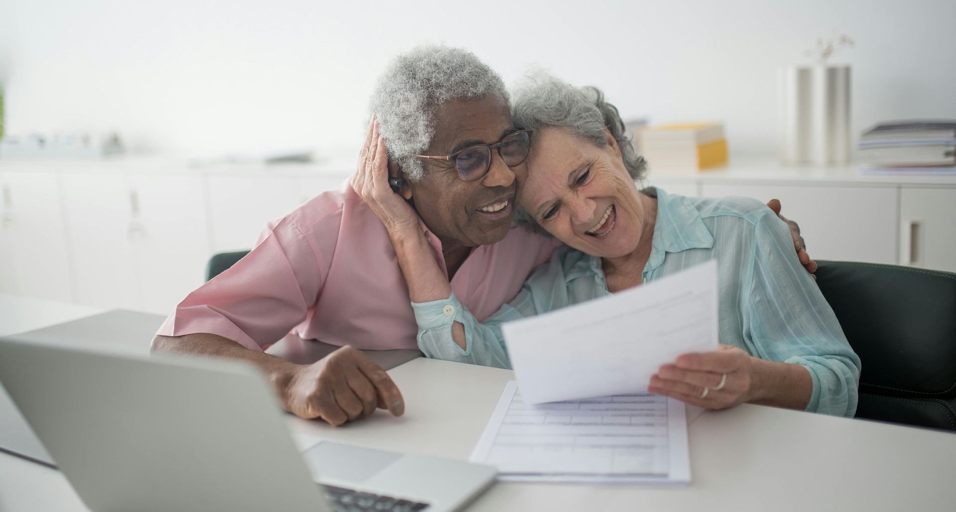 Elderly couple smiles while reviewing paperwork at a desk with a laptop.
