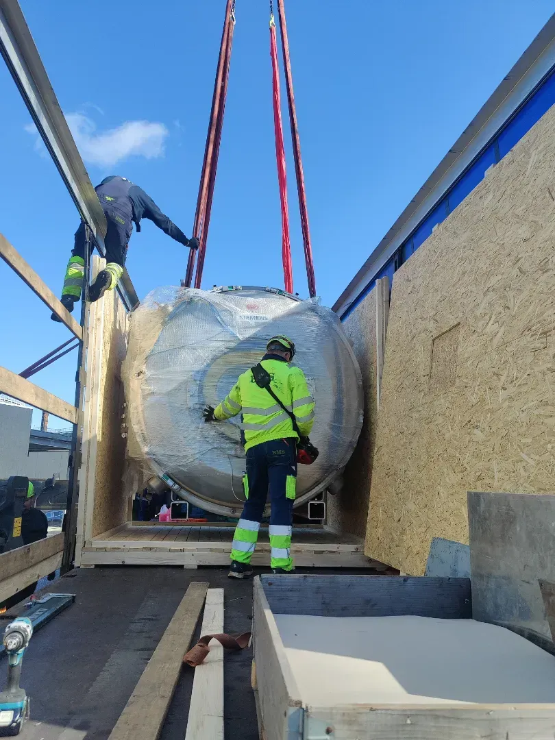 Arbejdere løfter en stor, indpakket cylindrisk genstand med stropper ned på et lad.