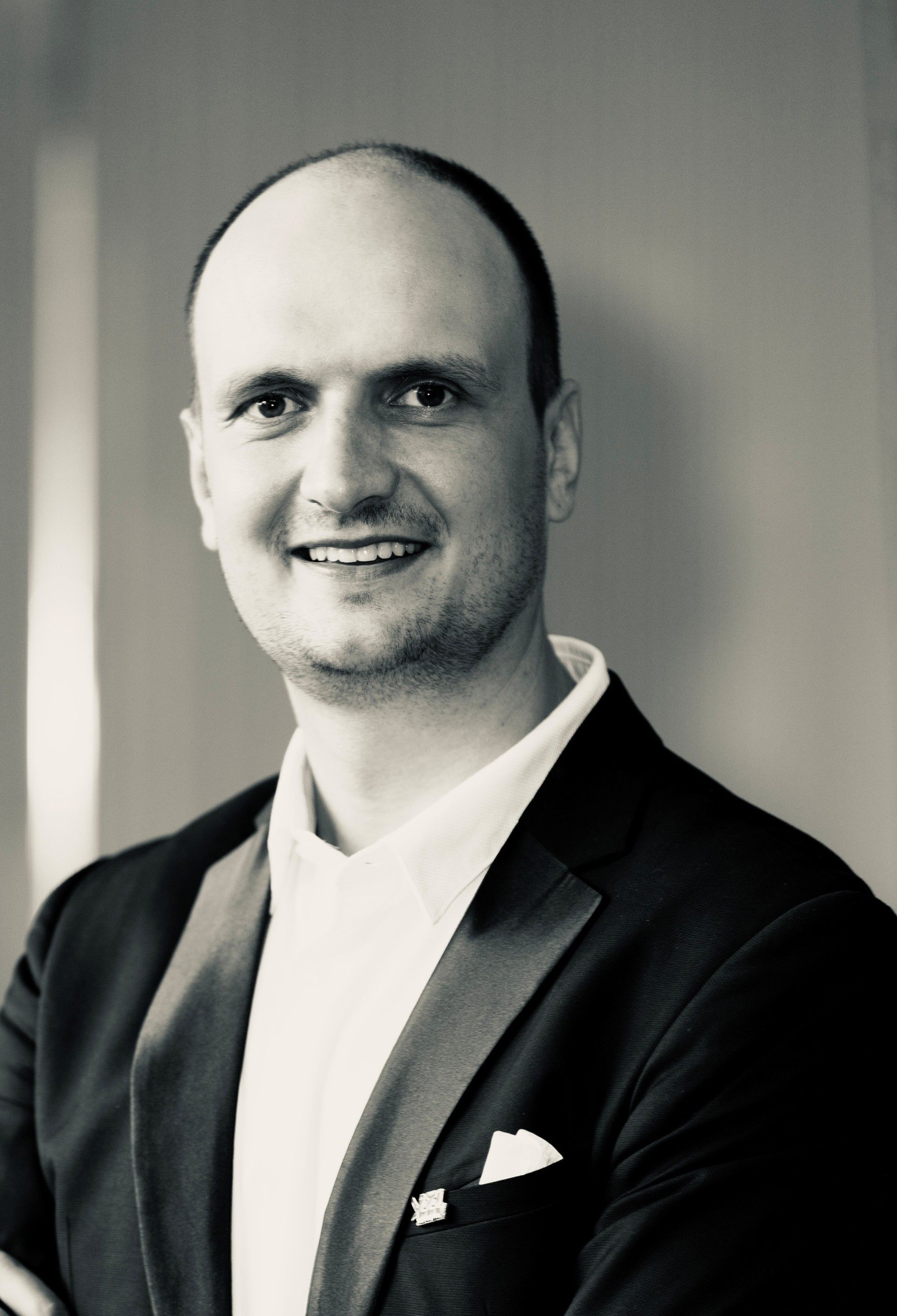 Portrait of a smiling man in a dark blazer and white shirt against a plain gray background