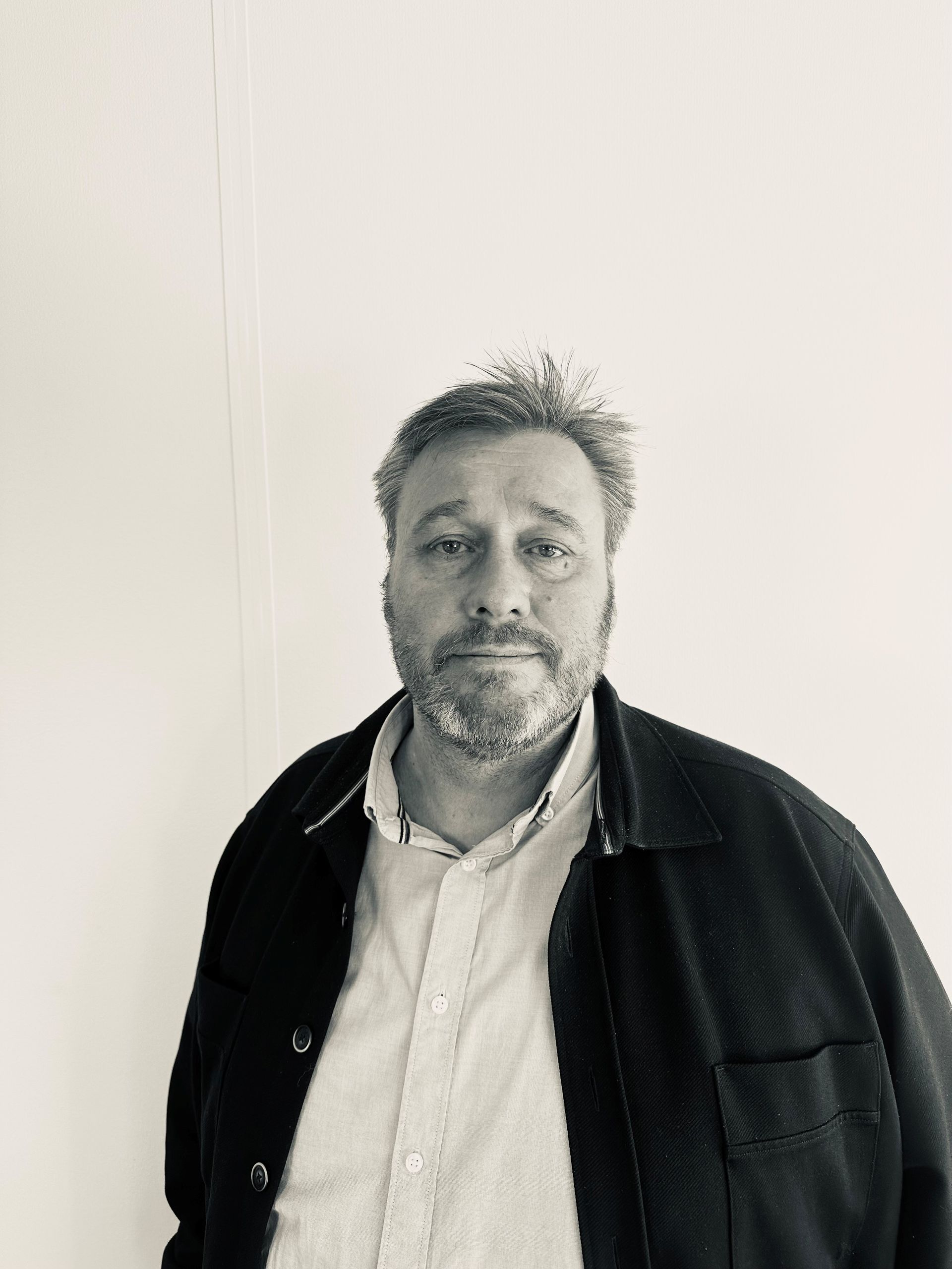 Man standing against a plain white wall, wearing a dark jacket over a light button-up shirt.
